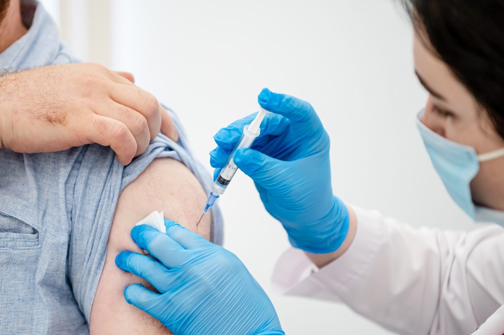 The image features a man with his arm extended, receiving an injection from a woman who is wearing blue gloves and a white shirt. She appears to be a nurse or medical professional attending to the patient. The man's arm is resting on a table, which occupies most of the background.nnIn addition to the main subjects, there are two other people in the scene: one person located near the left edge and another person at the far right side of the image. A bottle can be seen placed close to the center-right of the scene, possibly containing a medication or solution for the patient's treatment.