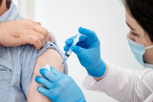The image features a man with his arm extended, receiving an injection from a woman who is wearing blue gloves and a white shirt. She appears to be a nurse or medical professional attending to the patient. The man's arm is resting on a table, which occupies most of the background.nnIn addition to the main subjects, there are two other people in the scene: one person located near the left edge and another person at the far right side of the image. A bottle can be seen placed close to the center-right of the scene, possibly containing a medication or solution for the patient's treatment.